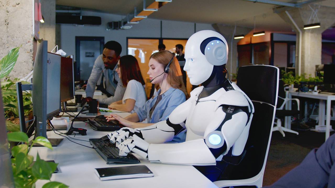 robot al lavoro in un ufficio, seduto alla scrivania insieme ad altri colleghi umani